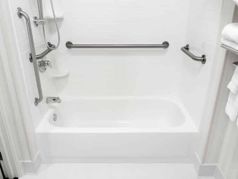 Baignoire blanche et douche accessible avec barres d'appui chromées, carrelage mural blanc et pommeau de douche amovible.