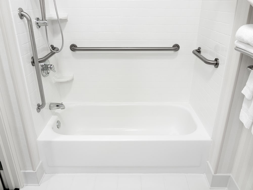 Baignoire blanche et douche accessible avec barres d'appui chromées, carrelage mural blanc et pommeau de douche amovible.