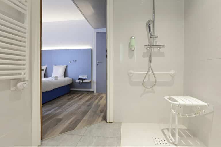 Douche accessible PMR avec siège rabattable et barres d'appui, s'ouvrant sur une chambre d'hôtel moderne avec lit bleu, tête de lit éclairée et parquet.