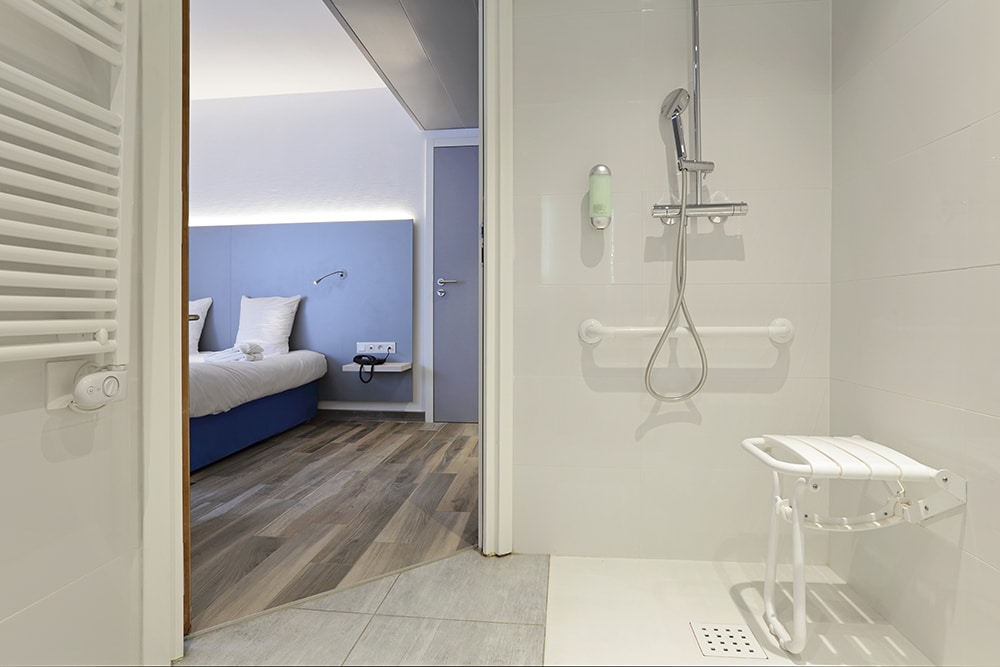 Douche accessible PMR avec siège rabattable et barres d'appui, s'ouvrant sur une chambre d'hôtel moderne avec lit bleu, tête de lit éclairée et parquet.