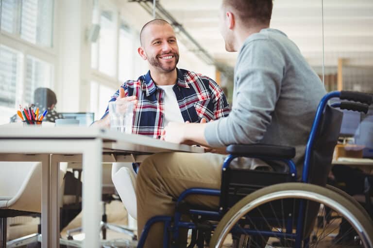 Deux hommes souriants discutent à une table de bureau. L'un est chauve et gesticule, l'autre est en fauteuil roulant.