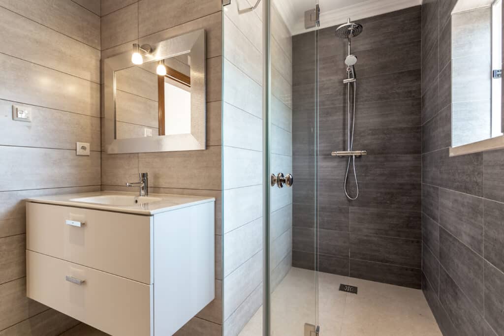 Salle de bain moderne : lavabo, douche à l'italienne & miroir Salle de bain moderne carrelée beige et gris. Lavabo suspendu blanc, miroir argenté, douche vitrée avec pommeau pluie chromé et mélangeur.